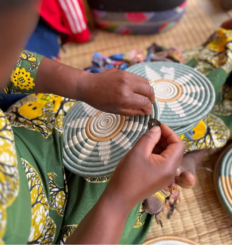 A close-up of a person's hands weaving a round woven placemat with concentric circles of sage green, white, and tan. The person is wearing a green dress with a yellow and black pattern, sitting on a woven mat floor with other woven items in the background.